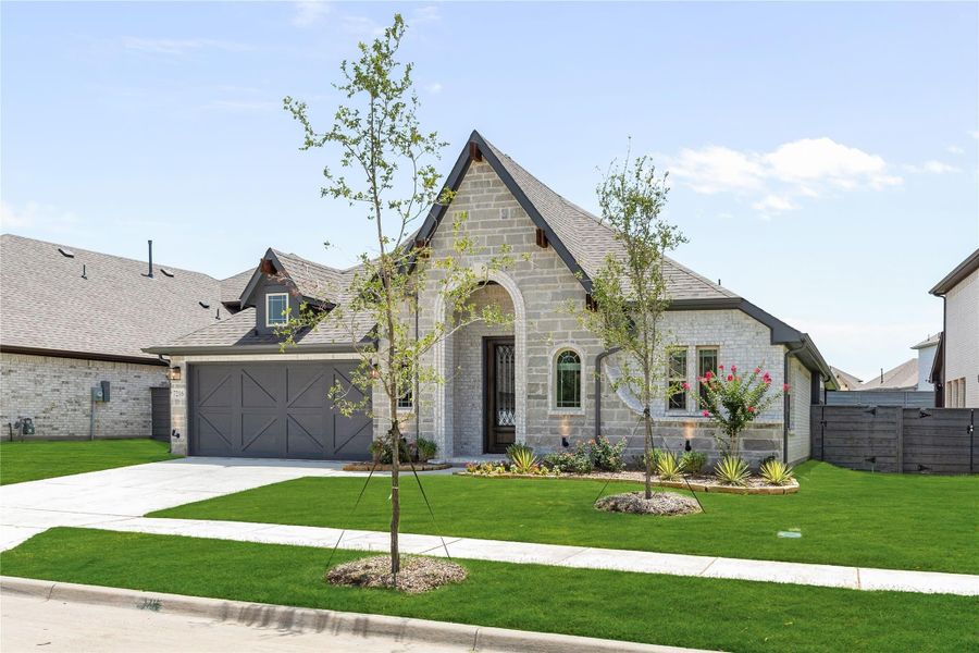 Single-story stone home with arched entry, two-car garage, and landscaped front yard with young trees Single-story stone home with arched entry, two-car garage, and landscaped front yard with young trees