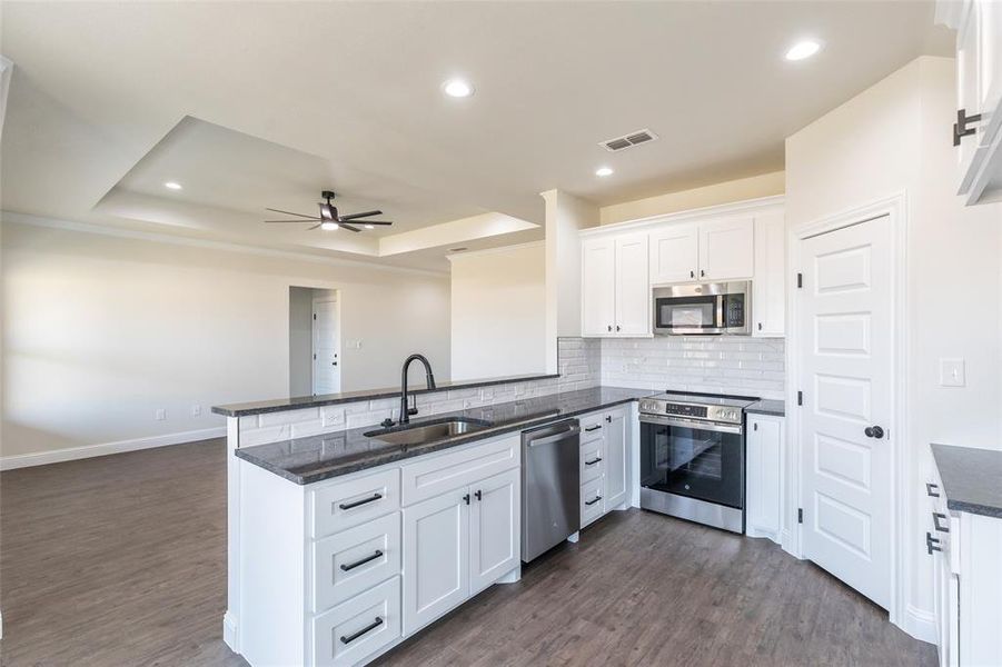 Modern kitchen featuring white shaker cabinetry, stainless steel appliances, dark countertops, a subway tile backsplash, and a large island with an undermount sink and matte black faucet