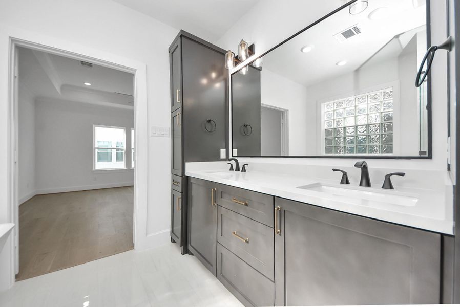 A contemporary bathroom with a double vanity featuring dark cabinetry and a large mirror. Glass block windows provide natural light and privacy, while the layout opens seamlessly to a spacious, light-filled bedroom.