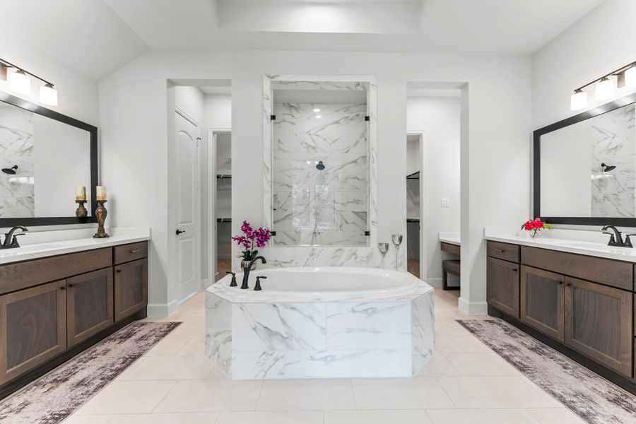 This is a wide shot of a luxurious primary bathroom. It features two dark wood vanities with white countertops and large, black-framed mirrors on either side. In the center is a white, marble-paneled bathtub, and behind it, a glass-enclosed shower with white marble-style tiles. This is a wide shot of a luxurious primary bathroom. It features two dark wood vanities with white countertops and large, black-framed mirrors on either side. In the center is a white, marble-paneled bathtub, and behind it, a glass-enclosed shower with white marble-style tiles.