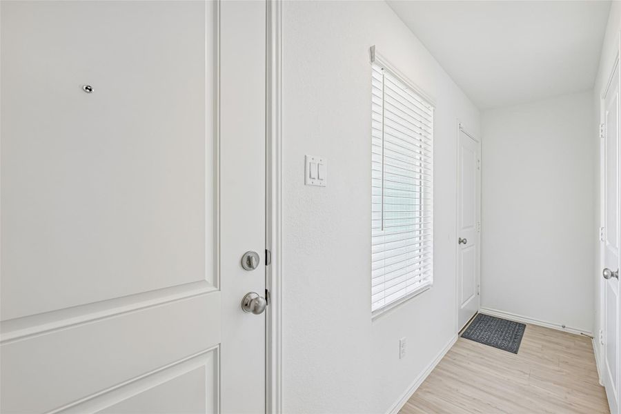 Foyer entry featuring a paneled door with brushed nickel hardware, light wood-finish flooring, a window with blinds, and white walls Foyer entry featuring a paneled door with brushed nickel hardware, light wood-finish flooring, a window with blinds, and white walls