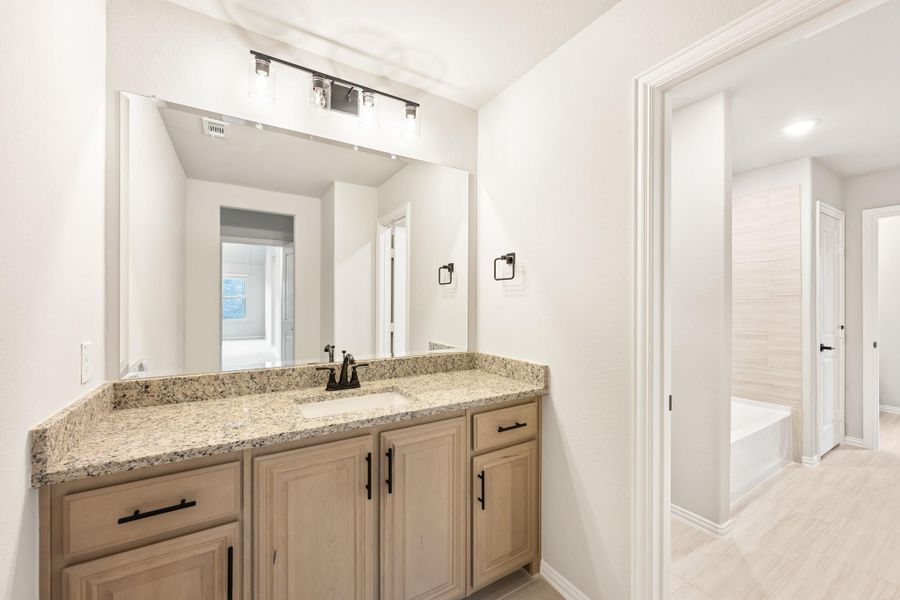 Bathroom vanity with granite countertop, double sinks, light wood cabinets, and large mirror with soaking tub visible in background. Bathroom vanity with granite countertop, double sinks, light wood cabinets, and large mirror with soaking tub visible in background.