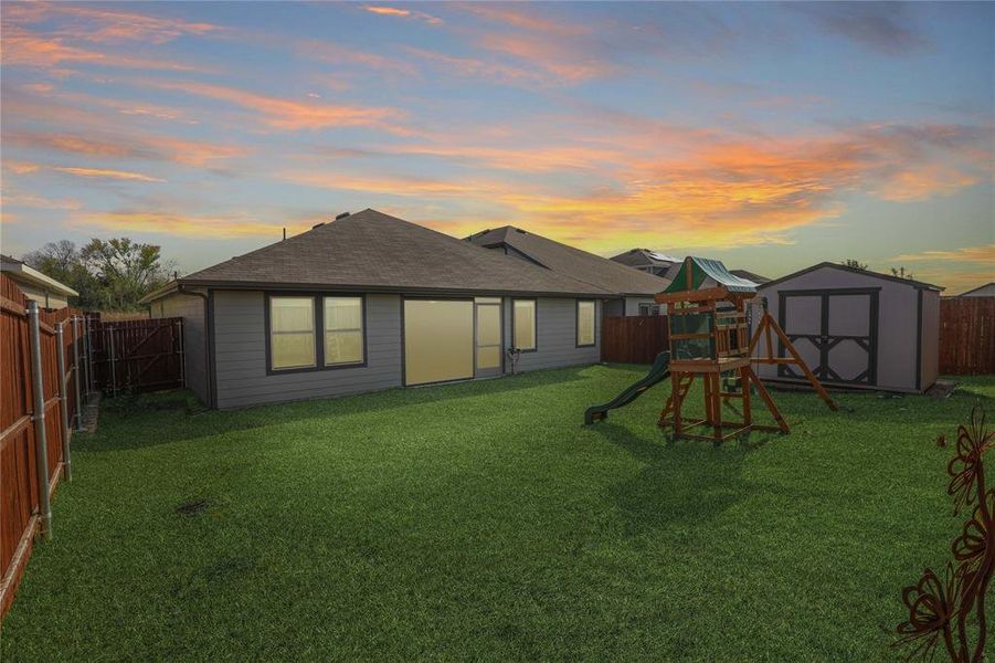 Back of property at dusk featuring a fenced backyard, a shingled roof, a playground, and a shed