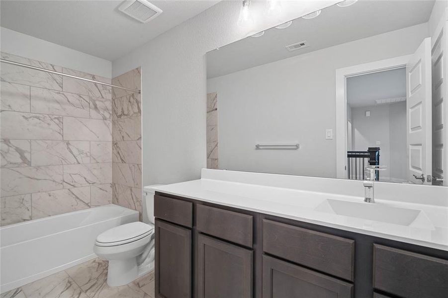 Secondary bath features tile flooring, bath/shower combo with tile surround, stained wood cabinets, beautiful light countertops, mirror, dark, sleek fixtures and modern finishes.