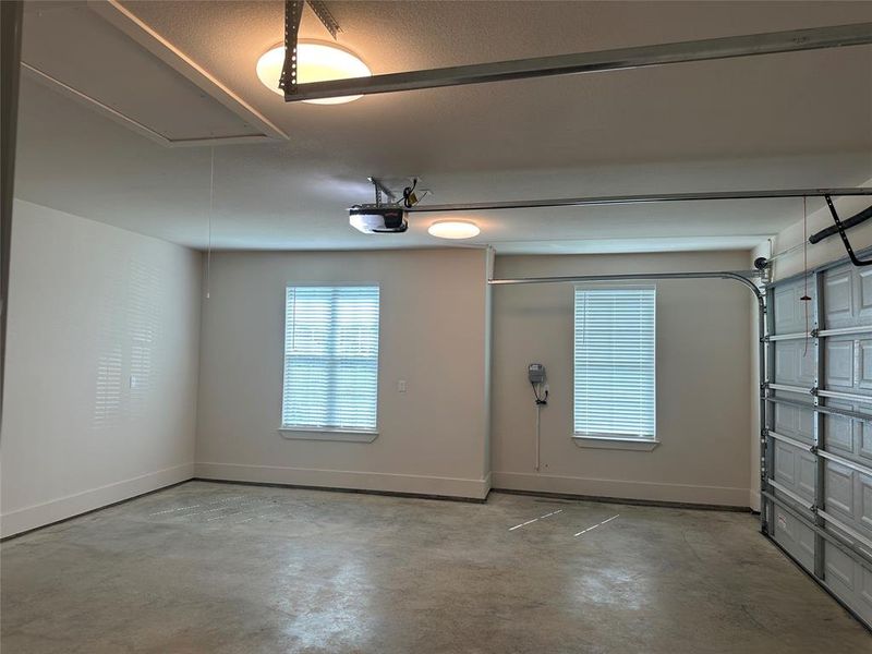 Spacious garage featuring a concrete floor, two windows with blinds, two ceiling-mounted light fixtures, a garage door opener, and an attic access panel