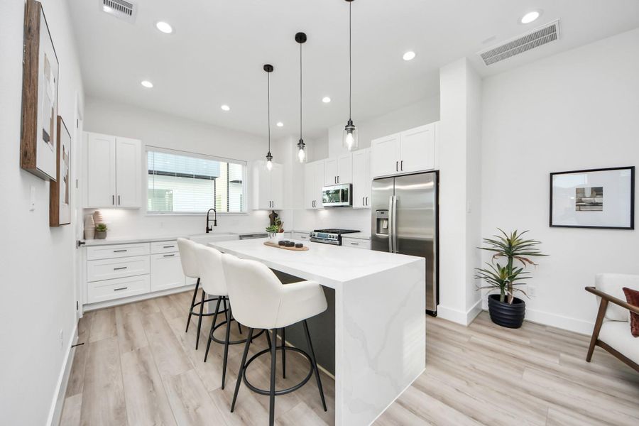 This modern kitchen features sleek white cabinetry, stainless steel appliances, and a spacious island with bar seating. The room is well-lit with recessed lighting and stylish pendant lights, complemented by light wood flooring for a fresh, airy feel.