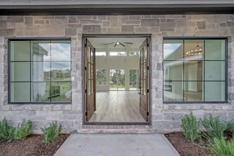 Grand double front doors open into a light-filled interior with soaring ceilings and expansive windows that frame peaceful views of the surrounding property.