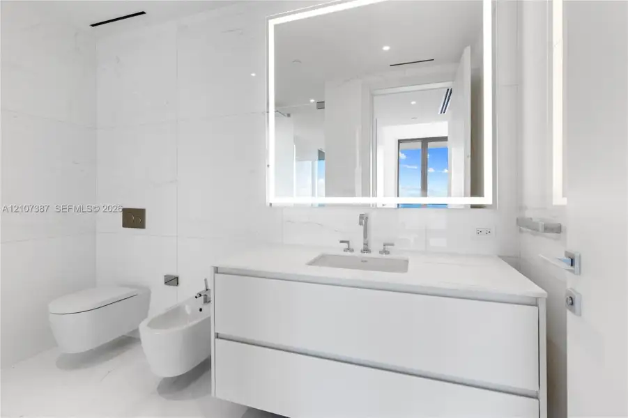 Modern floating vanity with a seamless countertop, wall-mounted commode and bidet, large-format tile wall cladding, and an integrated LED backlit mirror