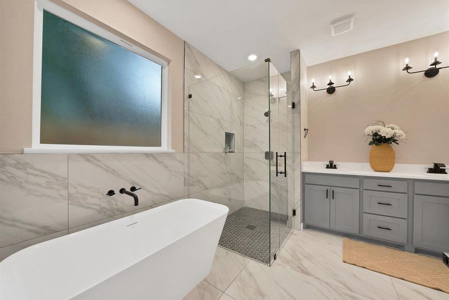 Contemporary bathroom featuring a freestanding tub with wall-mounted faucet, a glass-enclosed shower with marble-look tile surround, and dual vanities with gray cabinetry and black hardware Contemporary bathroom featuring a freestanding tub with wall-mounted faucet, a glass-enclosed shower with marble-look tile surround, and dual vanities with gray cabinetry and black hardware