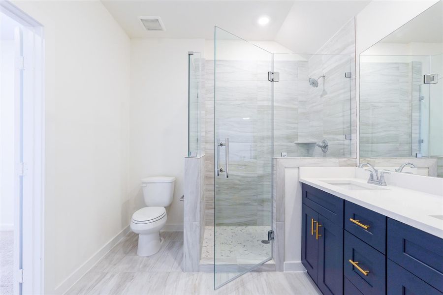 This bathroom features a sleek, modern design with a spacious glass-enclosed shower, a dual-sink vanity with bold blue cabinetry and gold hardware, and a clean, bright aesthetic.