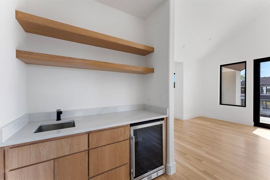 Indoor bar featuring open shelves, beverage cooler, light stone countertops, light wood finished floors, and light brown cabinets Indoor bar featuring open shelves, beverage cooler, light stone countertops, light wood finished floors, and light brown cabinets