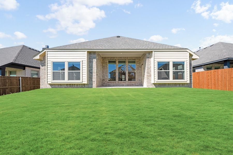 Rear exterior of single-story brick home with large windows, covered patio, and spacious green backyard with wood fencing.