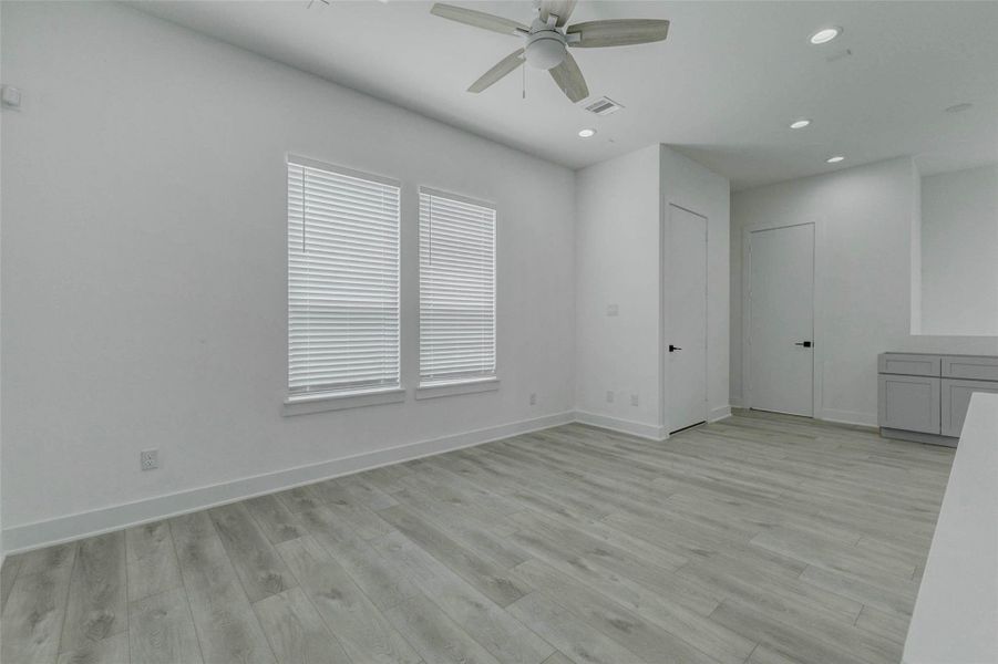 Bright living area with high ceilings, recessed lighting, ceiling fan, large windows with blinds, and seamless light wood-look flooring. The clean white walls and generous open floor plan create an airy, flexible space that can easily accommodate a living room, dining setup, or personalized entertaining area.