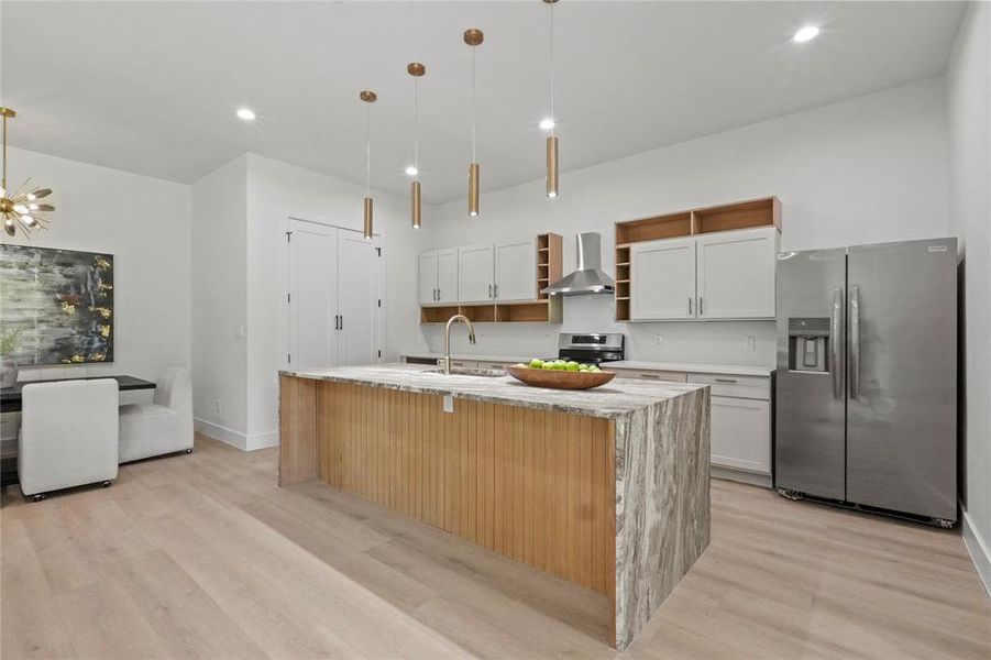 Kitchen with wall chimney range hood, light hardwood / wood-style flooring, a center island with sink, and stainless steel appliances