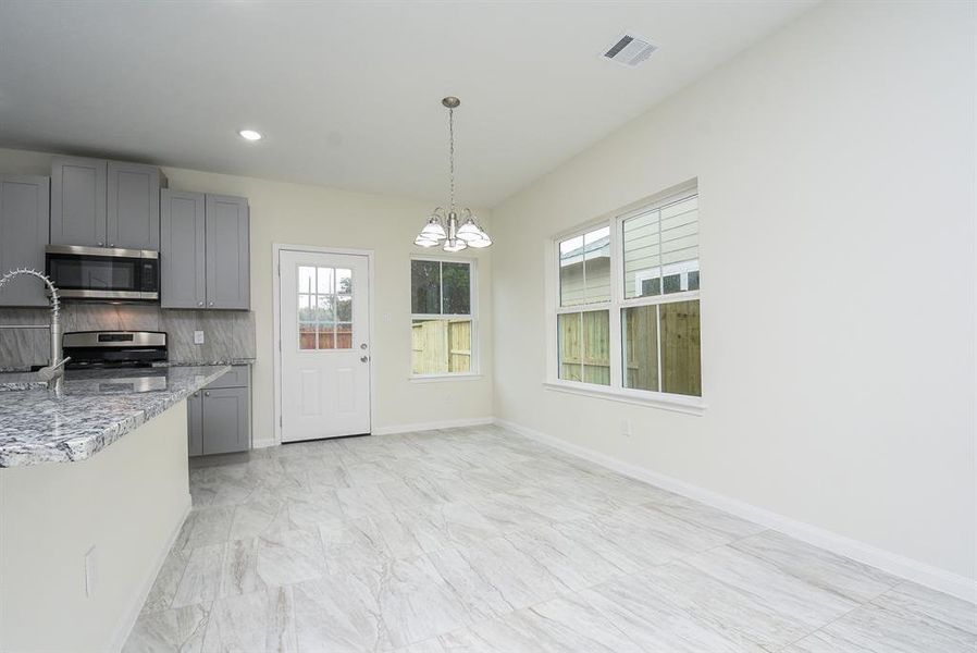 Modern kitchen with gray cabinets, stainless steel appliances, pendant lighting, and a view of a wooden fenced backyard through large windows.