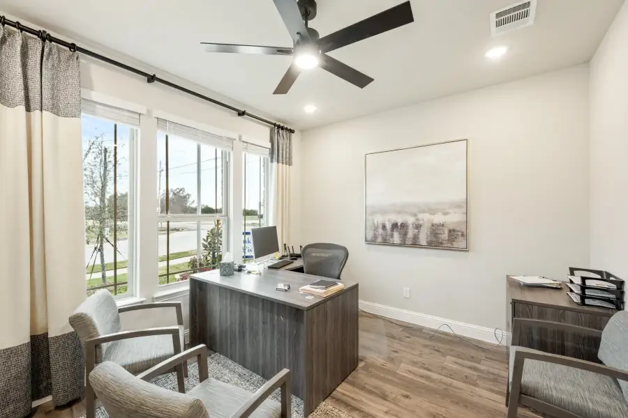 Home office with dark wood desk, ceiling fan, hardwood floors, and large windows with curtains