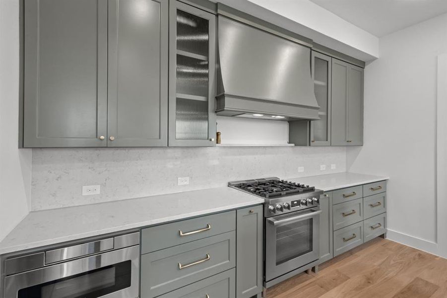 Kitchen with appliances with stainless steel finishes, wall chimney range hood, light stone countertops, light hardwood / wood-style floors, and backsplash