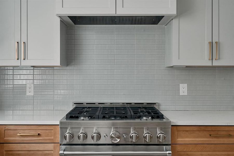 Stainless steel gas range with a multi-burner cooktop, light grey subway tile backsplash, white upper cabinetry with brass hardware, wood-finish lower cabinetry, and light-toned countertops Stainless steel gas range with a multi-burner cooktop, light grey subway tile backsplash, white upper cabinetry with brass hardware, wood-finish lower cabinetry, and light-toned countertops