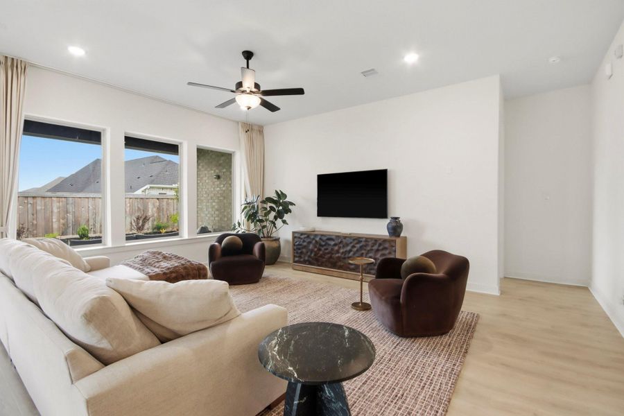 Sunlit living room highlighted by expansive windows, a statement ceiling fan, and tasteful furnishings, offering a comfortable space for relaxing and entertaining.