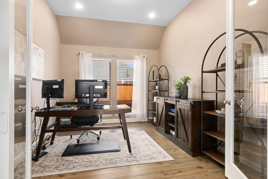 This photo shows a bright and inviting home office with glass French doors, hardwood flooring, and a vaulted ceiling, creating a functional workspace with natural light from a large window.