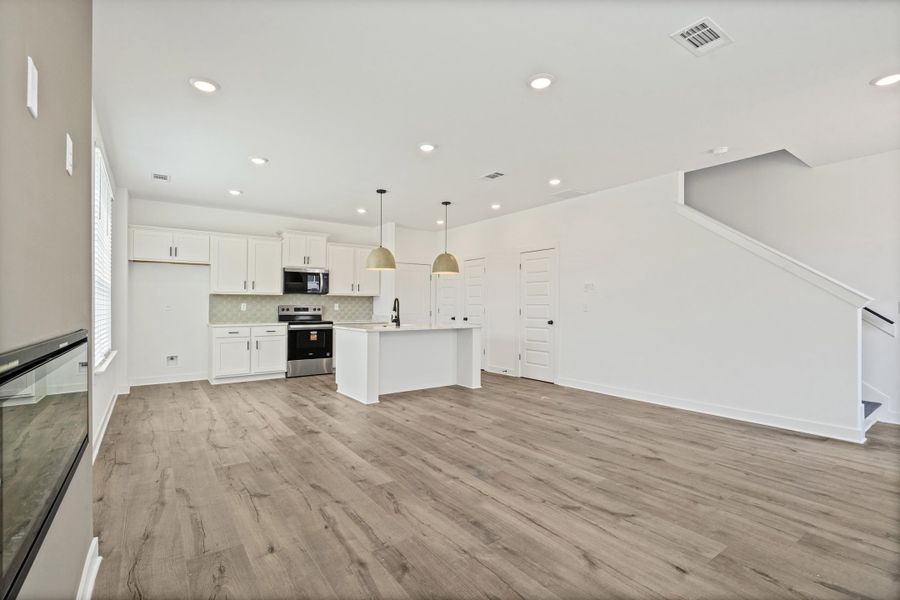 Open-concept living space with wood-finish flooring, recessed lighting, and a white kitchen featuring a large island with pendant lighting, white cabinetry, and stainless steel appliances