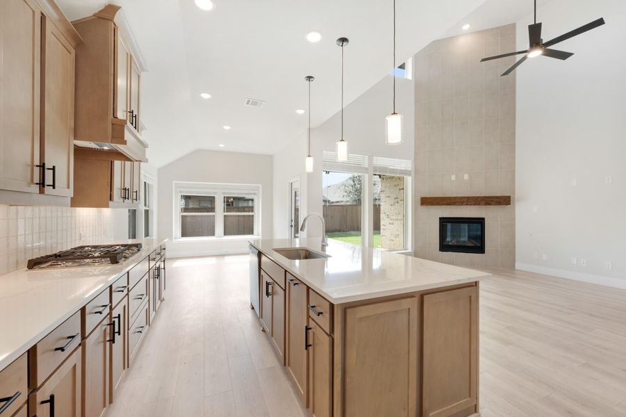 Open kitchen with large island, wood cabinets, gas cooktop, and vaulted ceiling open to living area with fireplace