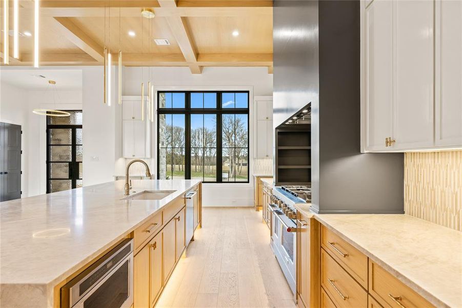 Kitchen featuring beamed ceiling, light stone countertops, a large island with sink, coffered ceiling, and light wood-type flooring