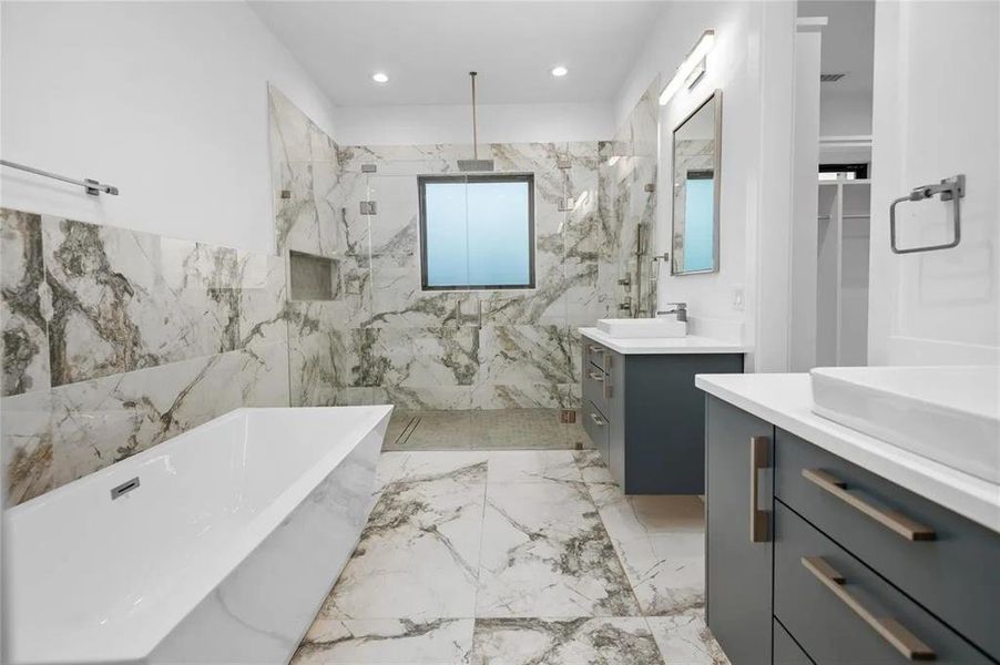 Spacious bathroom featuring large format marble-look tile flooring and walls, a freestanding soaking tub, a frameless glass shower enclosure, and dual vanity sinks with dark cabinetry Spacious bathroom featuring large format marble-look tile flooring and walls, a freestanding soaking tub, a frameless glass shower enclosure, and dual vanity sinks with dark cabinetry