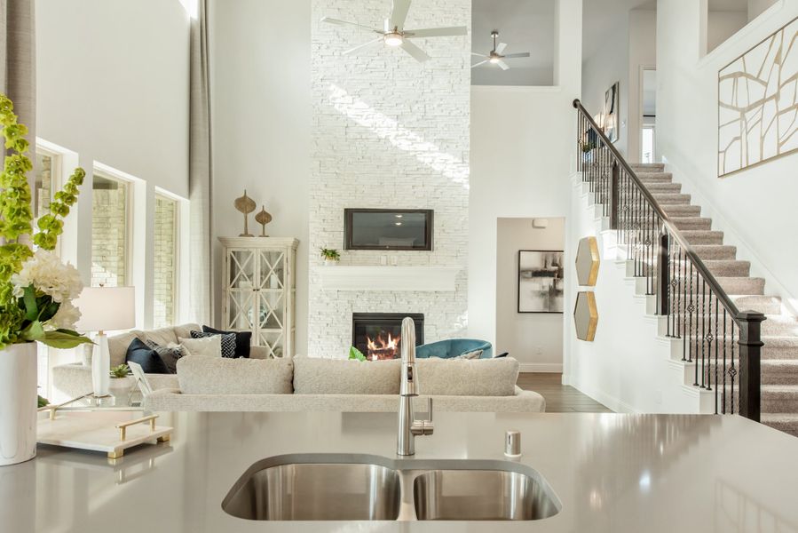 Open-concept home with kitchen sink in foreground, two-story living room, stone fireplace, and staircase