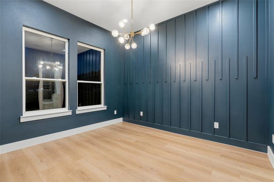 Room featuring a decorative accent wall with vertical paneling, two double-hung windows, wood-finish flooring, and a modern chandelier with spherical lights Room featuring a decorative accent wall with vertical paneling, two double-hung windows, wood-finish flooring, and a modern chandelier with spherical lights