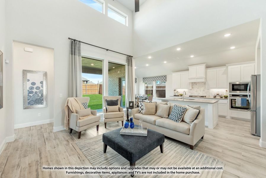 Open-concept living room with two-story ceilings, light wood floors, and view into white kitchen with stainless appliances.