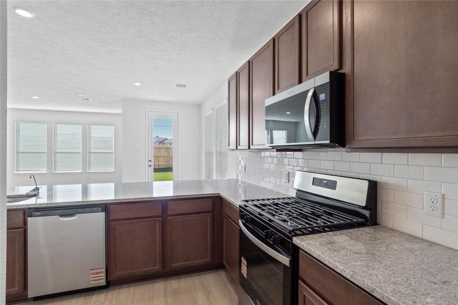 This spacious kitchen boasts soaring ceilings, Woodmont Cody cabinets in a Ginger stain, granite countertops, stainless steel Whirlpool appliances, a modern Daltile backsplash, recessed lighting, an extended breakfast bar countertop, and a pantry—all overlooking the expansive family room.