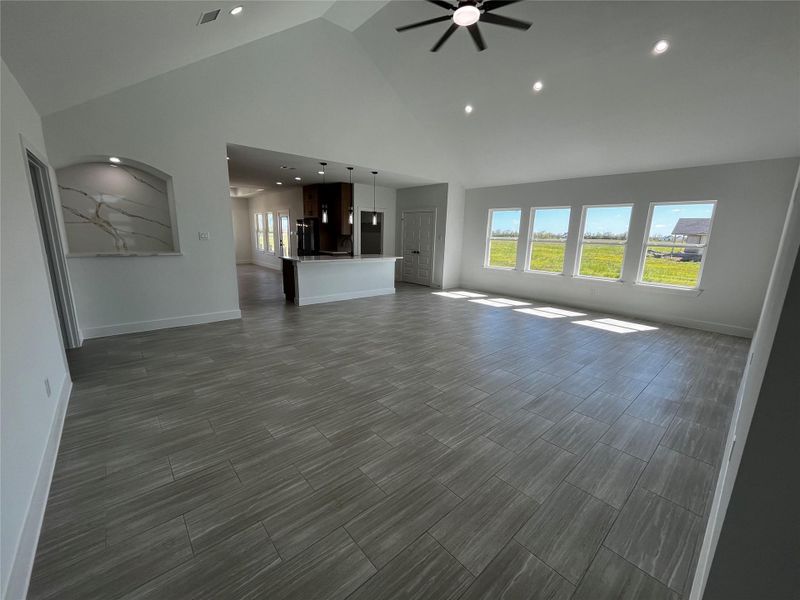 This spacious living area features high ceilings, large windows with ample natural light, modern tile flooring, and an open layout leading to a kitchen with a central island. Ideal for bright, airy living.