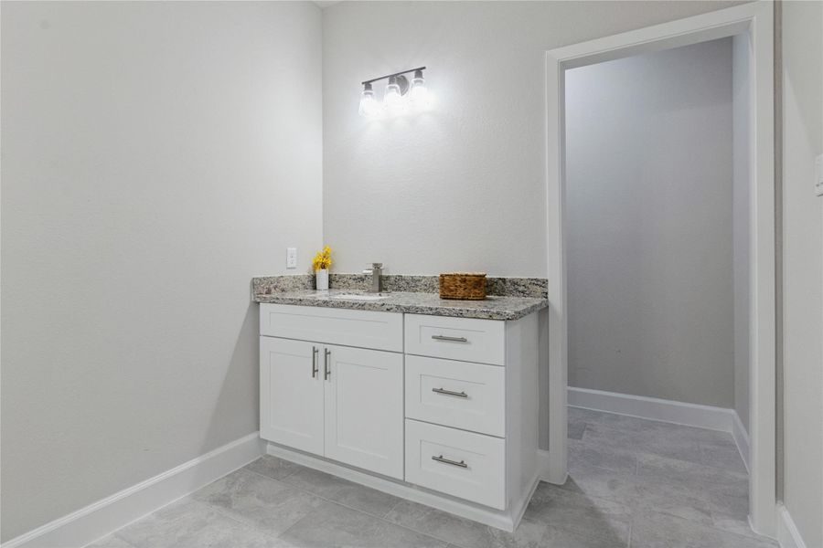 A modern bathroom with a sleek white vanity featuring a granite countertop and ample storage. The space is well-lit and has light gray tile flooring.