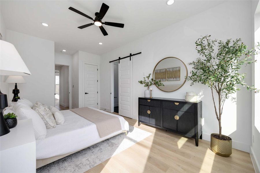 This light-filled primary bedroom features expansive windows, warm wood-look flooring, and elegant staging that highlights the room’s generous layout; a sleek black sideboard with contemporary décor anchors the space, while a modern ceiling fan and stylish sliding barn door lead seamlessly into the en-suite bathroom.