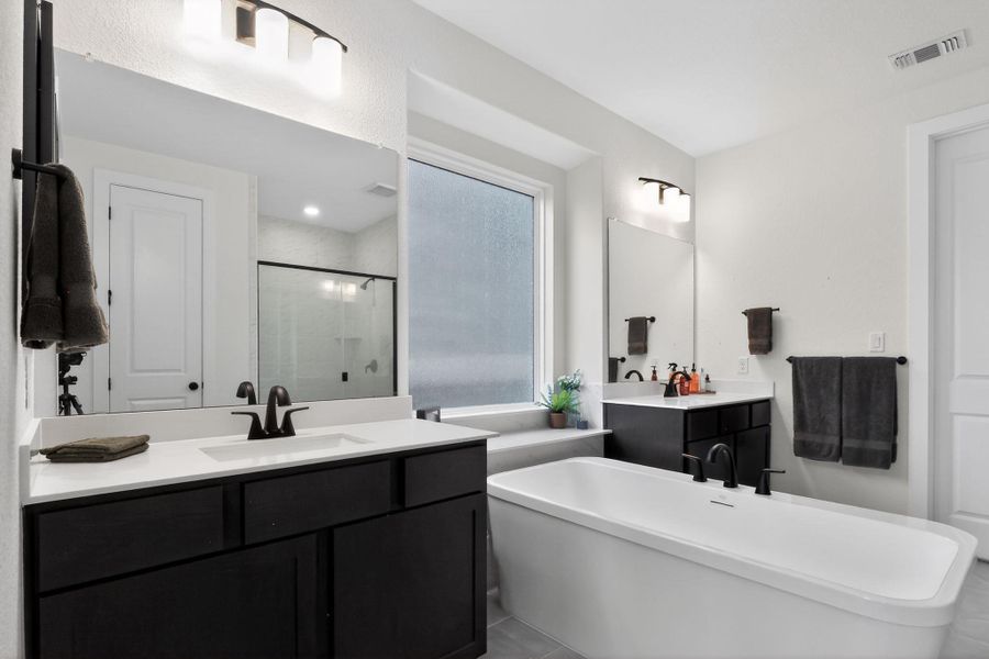 Freestanding soaking tub with black fixtures, dual vanities featuring dark cabinetry and white countertops, large wall-mounted mirrors, and an enclosed shower with a glass panel Freestanding soaking tub with black fixtures, dual vanities featuring dark cabinetry and white countertops, large wall-mounted mirrors, and an enclosed shower with a glass panel