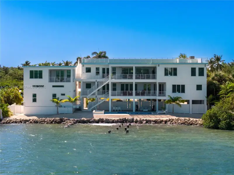 Multi-level waterfront structure featuring a white stucco exterior, tiered balconies with metal railings, an integrated ground-level swimming pool, and stone bulkhead shoreline reinforcement Multi-level waterfront structure featuring a white stucco exterior, tiered balconies with metal railings, an integrated ground-level swimming pool, and stone bulkhead shoreline reinforcement