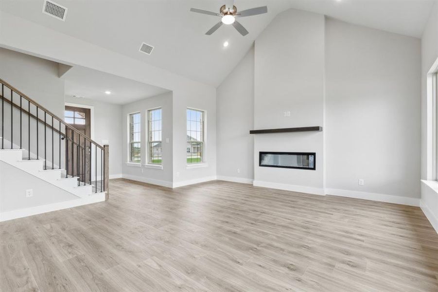 Unfurnished living room featuring high vaulted ceiling, ceiling fan, a glass covered fireplace, light wood-style flooring, and stairs Unfurnished living room featuring high vaulted ceiling, ceiling fan, a glass covered fireplace, light wood-style flooring, and stairs