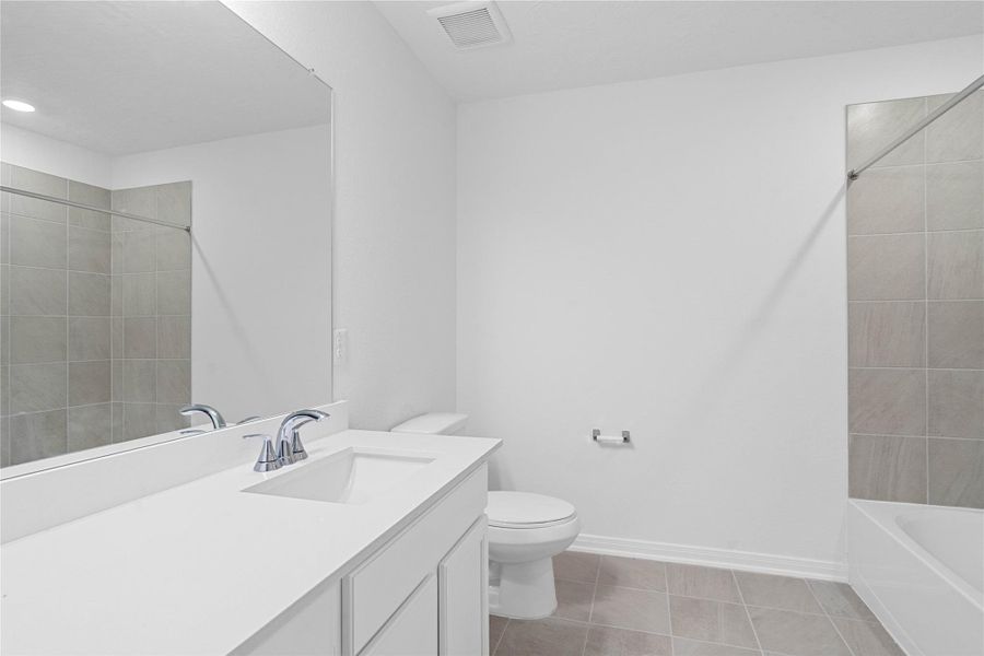 The secondary bathroom offers tile flooring, a bath/shower combination with a stylish tile surround, rich stained wood cabinetry with added knee space and elegant light countertops, a mirror, and sleek fixtures with modern finishes.