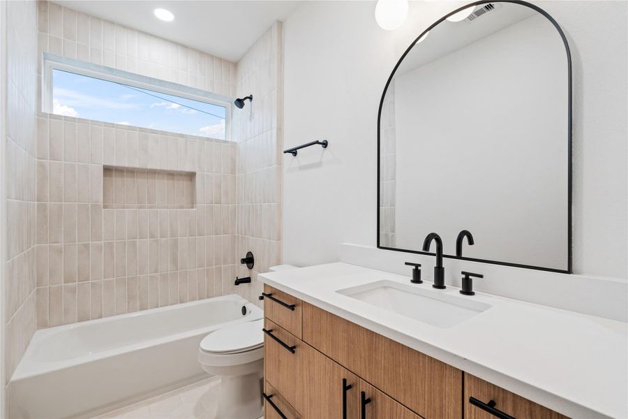 A beautifully appointed secondary bathroom with designer touches at every turn—featuring a floating wood vanity, quartz countertop, and a textured vertical tile surround with built-in niche. Matte black hardware and an arched mirror add a bold, modern edge to this serene and functional space.