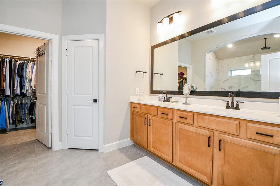 This photo features a spacious bathroom with a double vanity, large mirror, and ample cabinet space. It includes a walk-in closet with organized shelving and a glimpse of a modern shower area. The neutral tones and bright lighting create an inviting atmosphere.