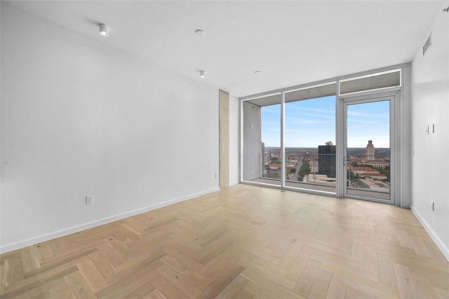 A minimalist design, featuring light-colored wood flooring arranged in a herringbone pattern. The room is brightly lit by natural light streaming in through large, floor-to-ceiling glass doors that lead to a balcony.