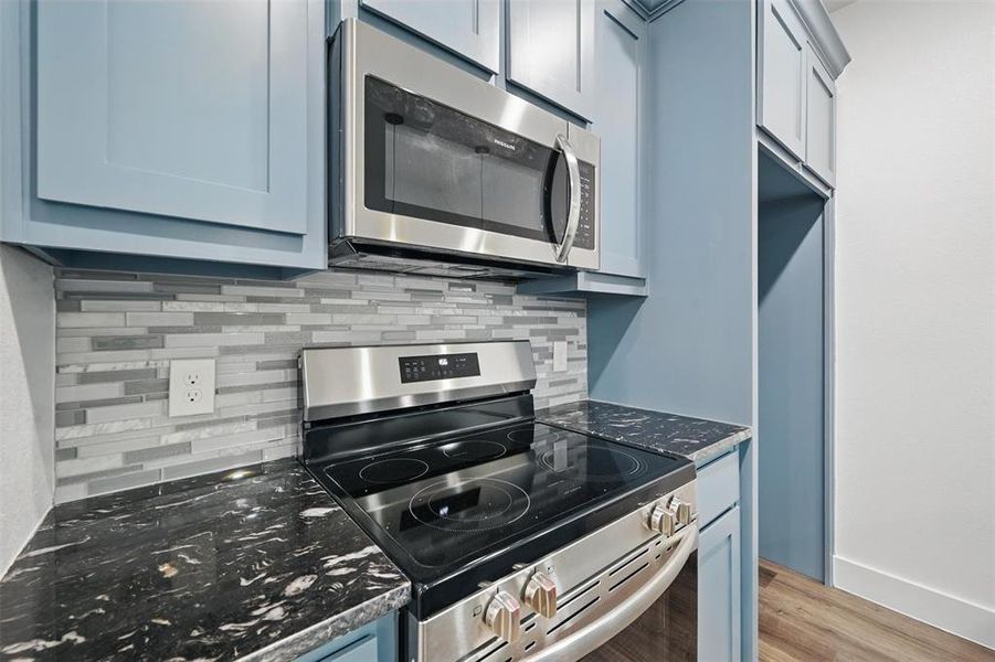 Kitchen featuring light wood-type flooring, dark stone countertops, blue cabinetry, decorative backsplash, and appliances with stainless steel finishes