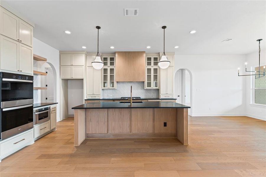 Kitchen featuring arched walkways, a center island with sink, stainless steel appliances, light wood-type flooring, and tasteful backsplash