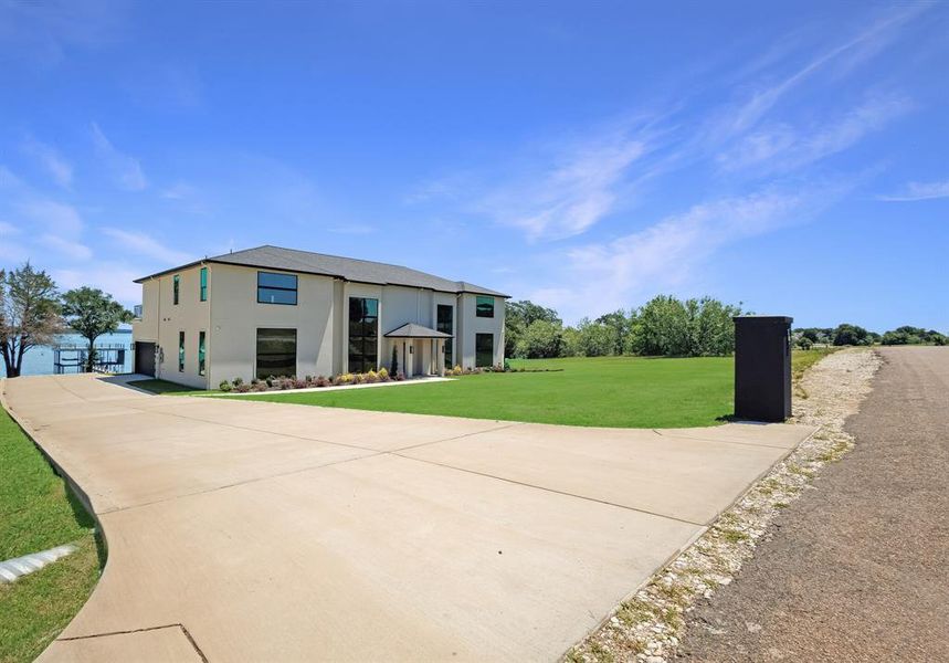 Contemporary stucco exterior with a multi-pane window design, a prominent front entrance with columns, a concrete driveway, a manicured lawn, and a waterfront setting with a private dock