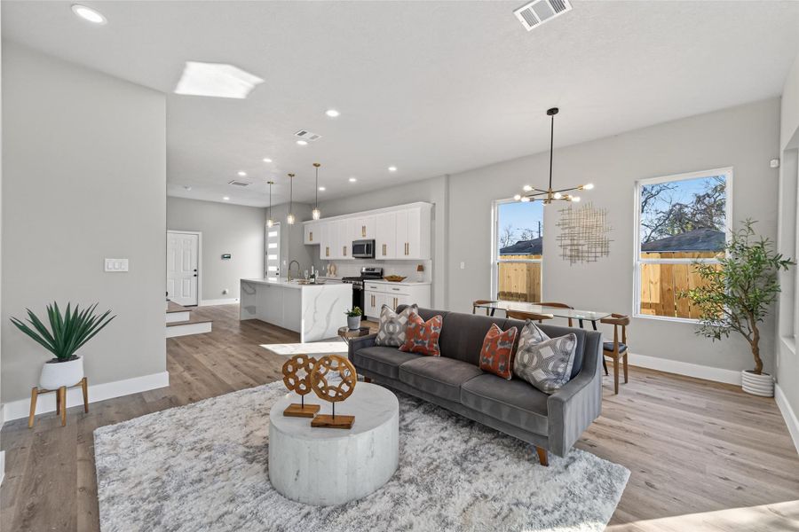 Open-concept living and dining area anchored by wide-plank white oak–finish laminate flooring, recessed LED lighting, and windows that flood the space with natural light.