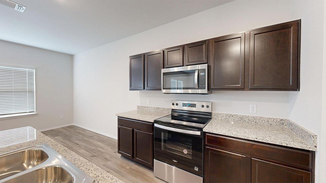 Kitchen with stainless steel appliances, dark wood finish cabinetry, light wood-type flooring, and light stone counters
