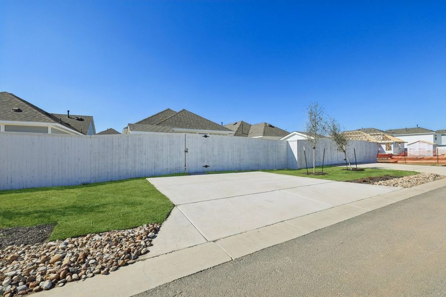 Image of a back lot with a white fence, cement driveway, green grass and a blue sky in the background