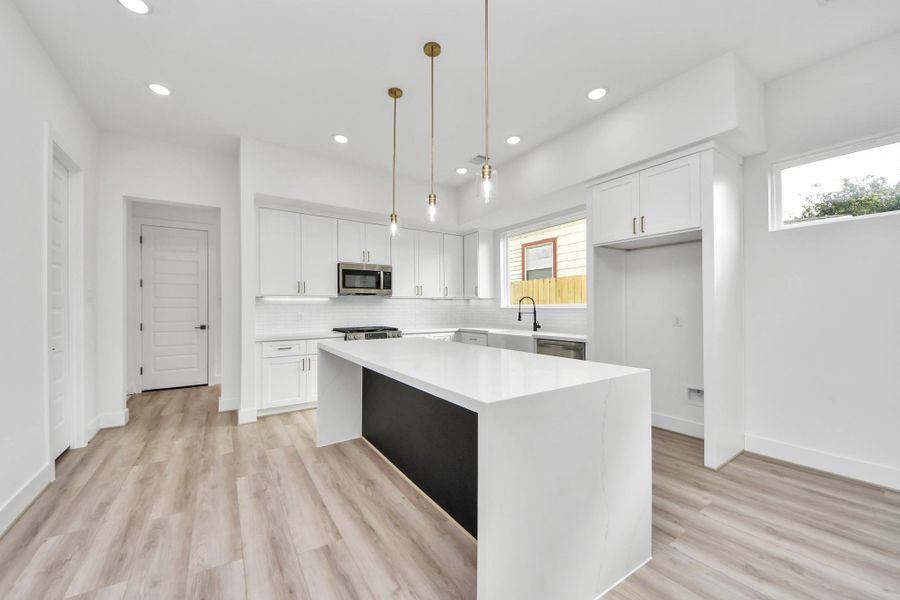 Bright, modern kitchen with sleek white cabinetry and a large island. It features stainless steel appliances, pendant lighting, and wood-style flooring, creating an inviting and functional space.
