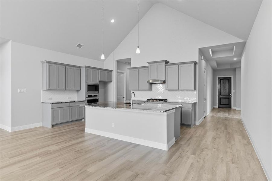 Kitchen featuring gray cabinets, backsplash, high vaulted ceiling, light stone counters, and an island with sink Kitchen featuring gray cabinets, backsplash, high vaulted ceiling, light stone counters, and an island with sink
